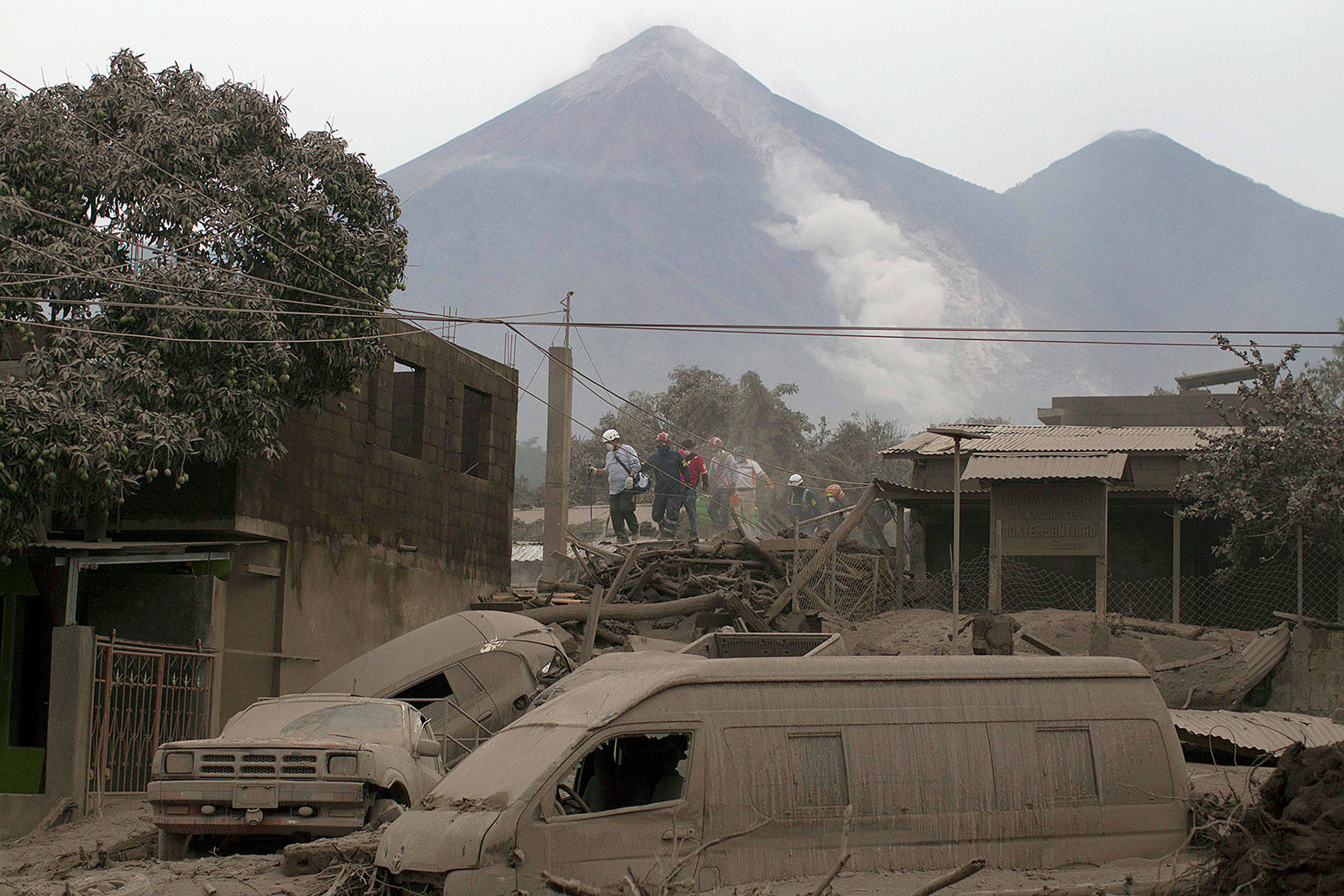 Several emergency responders wearing hard hats work amidst extensive destruction caused by a volcanic eruption. Ash covers everything, from collapsed buildings to multiple vehicles buried under debris. Two visible mountains loom in the background, one emitting a plume of smoke.
