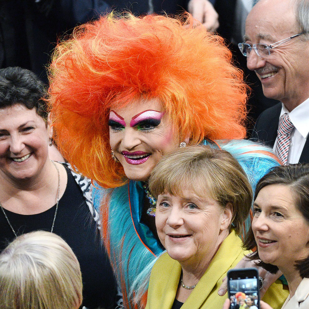 A group of five people posing for a photo at a social event. One person stands out with a vibrant orange wig and colorful makeup, while another person in a yellow jacket smiles at the camera. The group appears joyful in a lively gathering.
