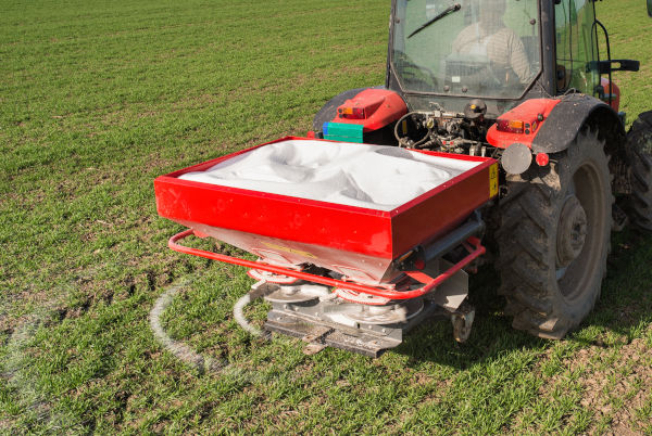 A red tractor equipped with a seed spreader parked on a green field, spreading white seeds or fertilizer under a clear sky.