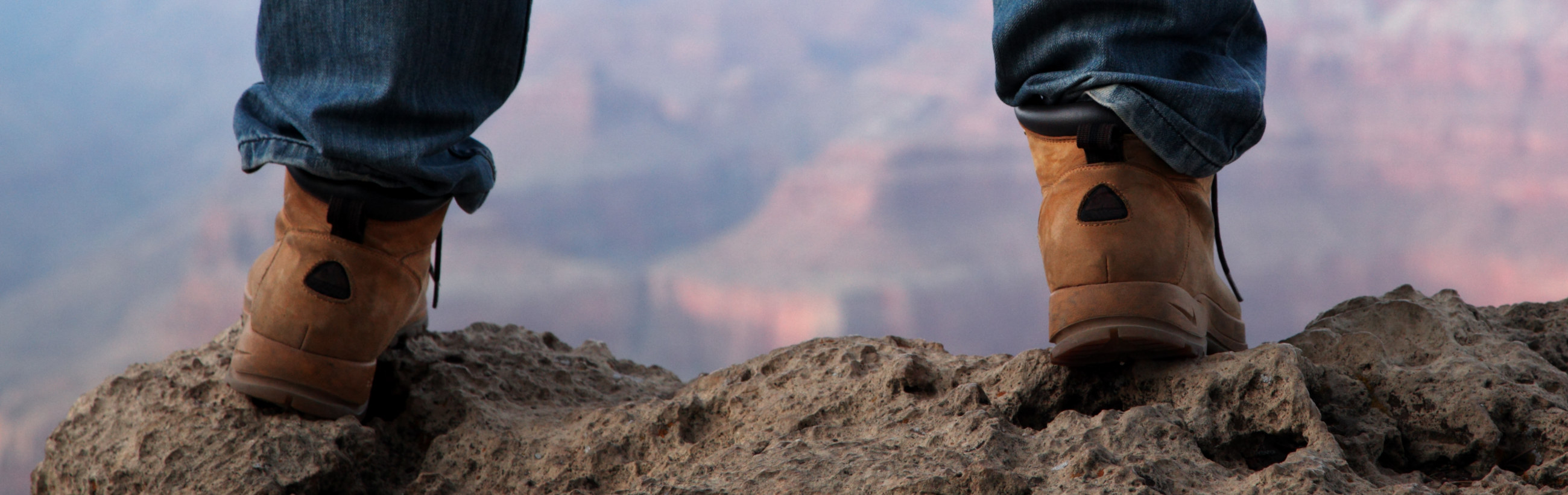 Close-up of a person's feet clad in worn tan boots, standing on a rocky outcrop with a blurred mountainous background bathed in soft, dusky light.