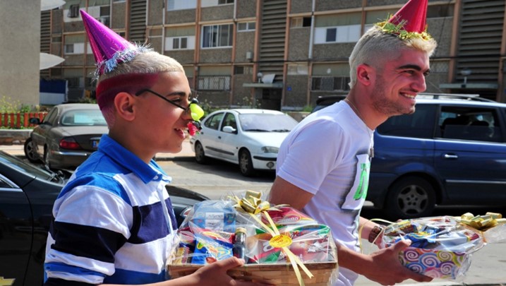 Two young men celebrating outdoors, wearing party hats and smiling brightly. One is holding several gift baskets. The setting includes a sunny day with a backdrop of apartment buildings and parked cars.