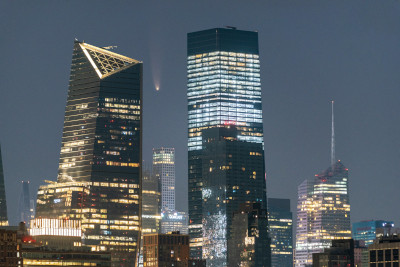 A city skyline at night featuring modern skyscrapers illuminated with lights under a clear sky, with a comet visible above the tallest buildings, symbolizing celestial phenomena.
