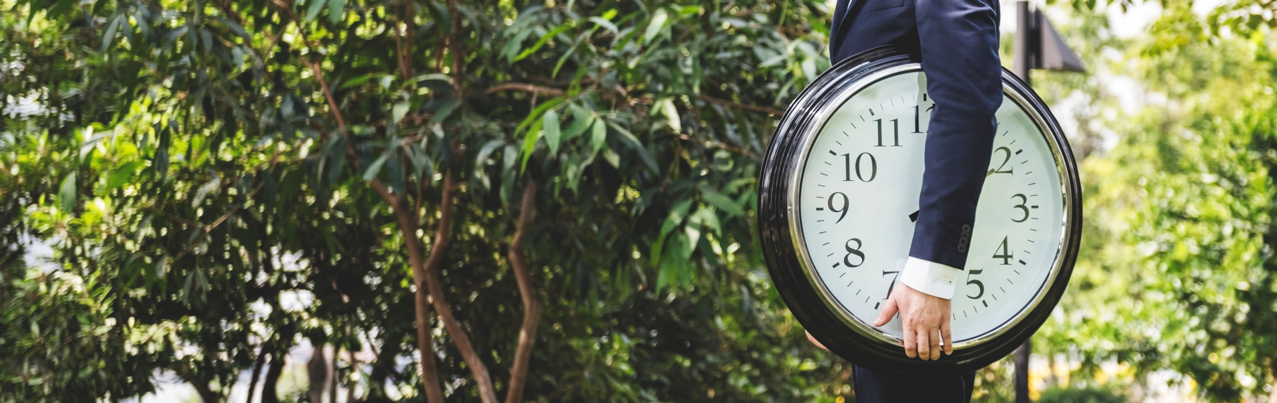 A person in a business suit stands with their head obscured by a large, round clock showing 10:10, set against a backdrop of dense, lush greenery.