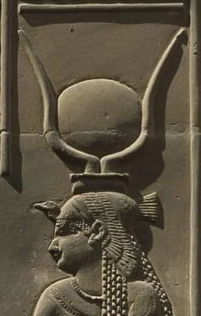 Bas-relief carving of an ancient Egyptian figure with an ornate headdress featuring a circular disk between two large, upward-curved horns. The figure is sculpted in profile, displaying detailed facial features and a stylized hair braid.