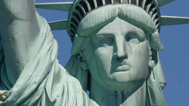 Close-up view of the Statue of Liberty's head and upper torso, highlighting her solemn expression and the prominent crown resembling rays of light. The sky in the background is a clear blue, emphasizing the statue's symbolic presence.