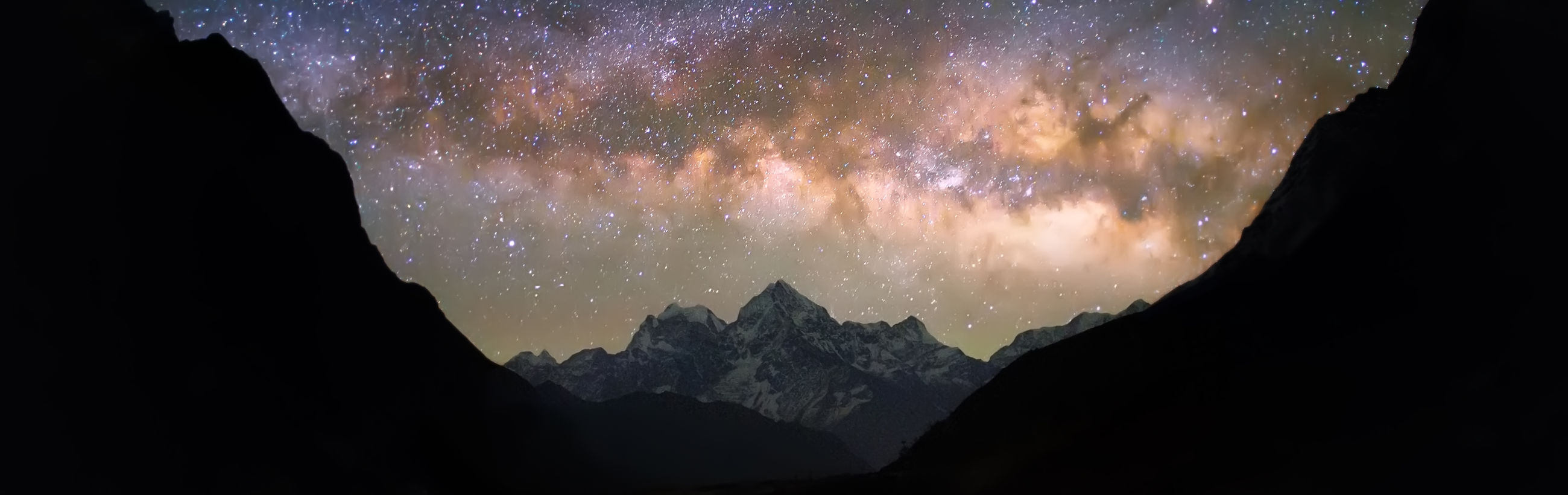 A panoramic view of a star-filled sky sprawling above snow-capped mountain ridges at night, with silhouetted rocky peaks framing the scene on either side.
