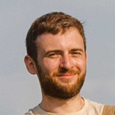 Portrait of a smiling young man with short brown hair and a beard against a clear sky backdrop.