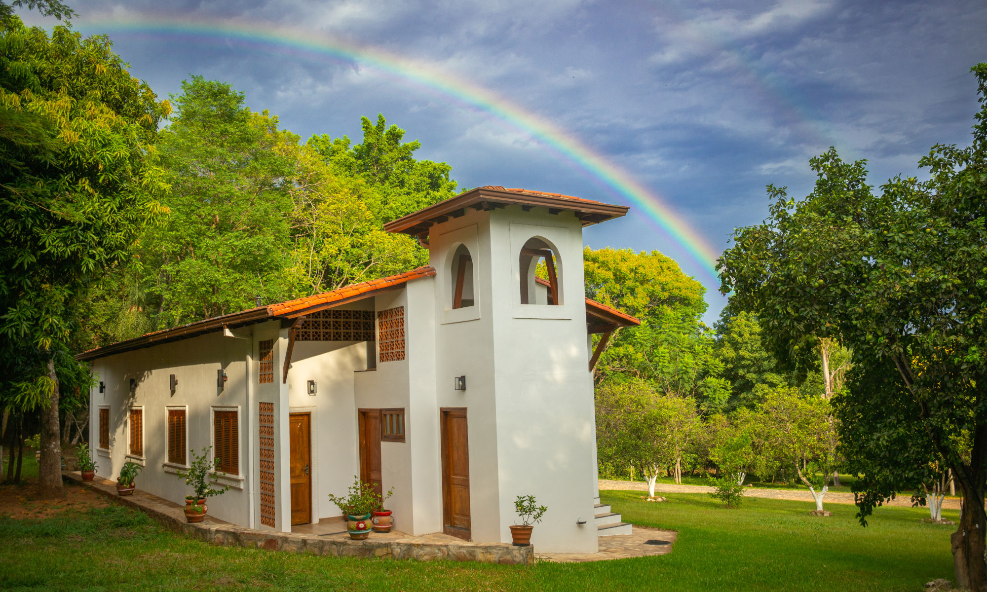 A picturesque white stucco house with terracotta tiled roof and wooden accents surrounded by lush greenery under a bright sky, with a vivid double rainbow arching overhead.