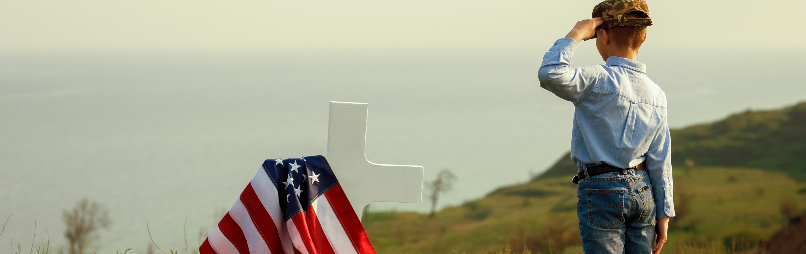 A man in casual attire and a camouflage cap stands on a grassy hill, gazing into the distance with one hand shielding his eyes from the sun. Beside him is a prominently displayed American flag draped over a white cross, suggestive of remembrance or tribute.