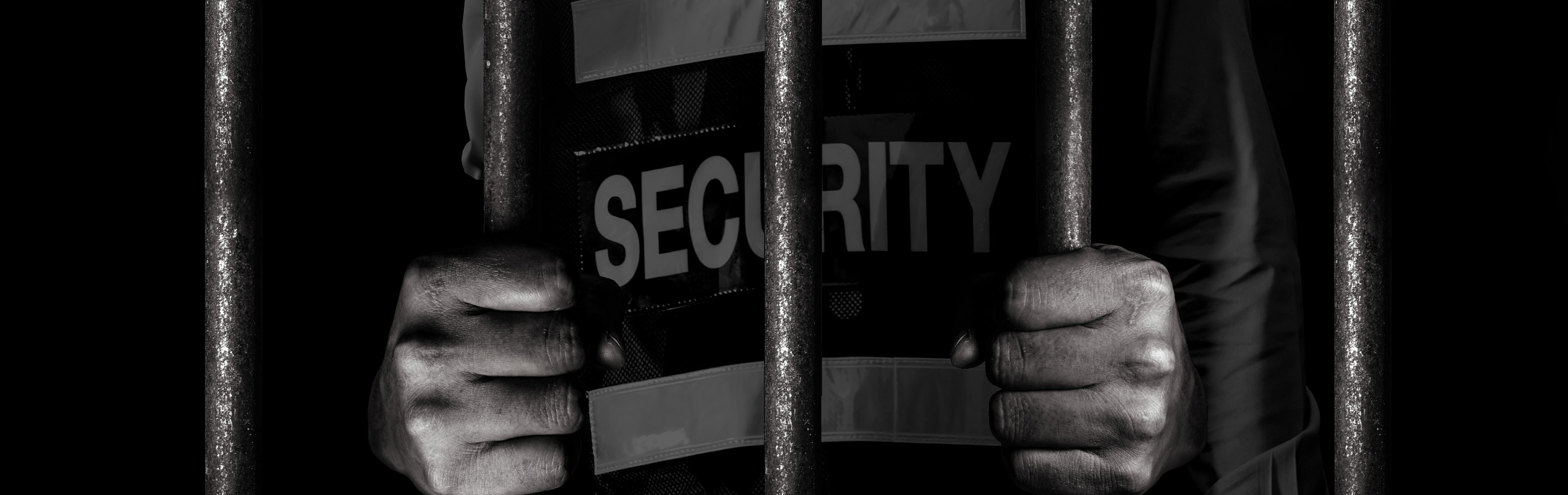 A grayscale image focusing on the hands of a person enclosed behind bars; the hands grasp the bars firmly and partially cover the word "SECURITY" printed on the person's attire.