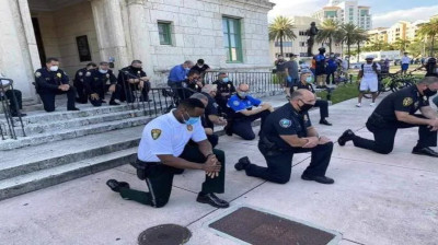 A group of law enforcement officers in uniform, including one wearing a white shirt and others in dark uniforms, kneel on one knee on the steps in front of a building. A diverse group of officers and public spectators, including some standing in the background under clear skies.