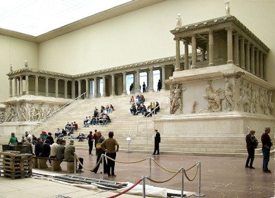 A detailed recreation of the Pergamon Altar in a museum setting, showcasing a large stepped platform and intricately carved friezes depicting titanic struggles and mythic tales. Visitors are scattered around the exhibit, observing and discussing the historical artifact in an expansive, well-lit hall.
