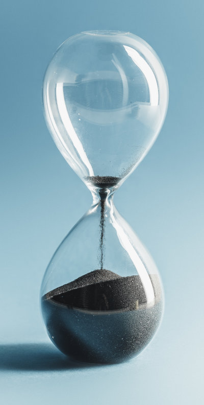 A clear glass hourglass with sand flowing from the top chamber to the bottom against a pale blue background, symbolizing the passage of time.