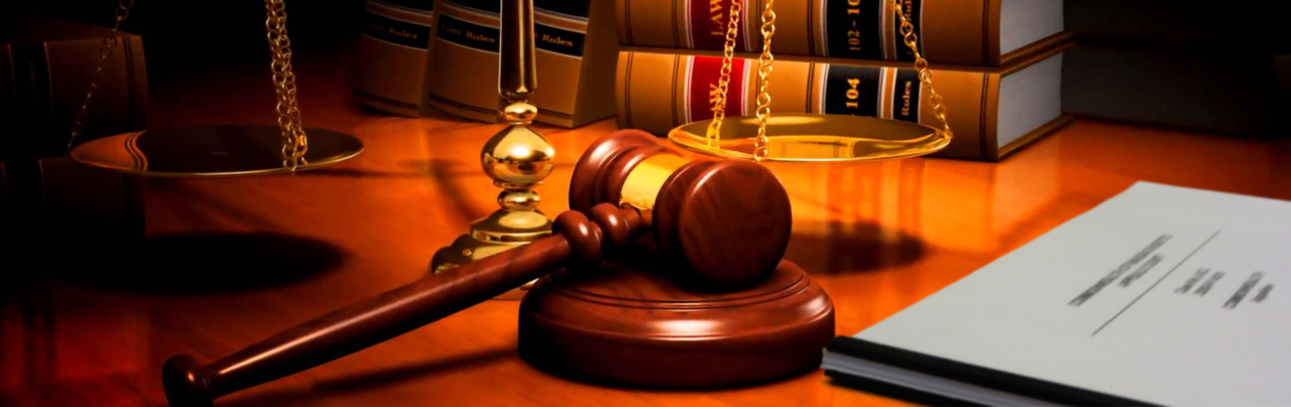 A polished wooden judge's gavel rests on its sound block, in front of a background featuring a golden scale balance, leather-bound law books, and a wooden cabinet. The scene is warmly lit, creating an atmosphere of judicial authority and solemnity.