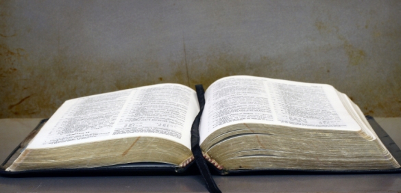 An open book with worn pages lies on a gray table, softly illuminated from above, displaying two visible pages filled with dense text.