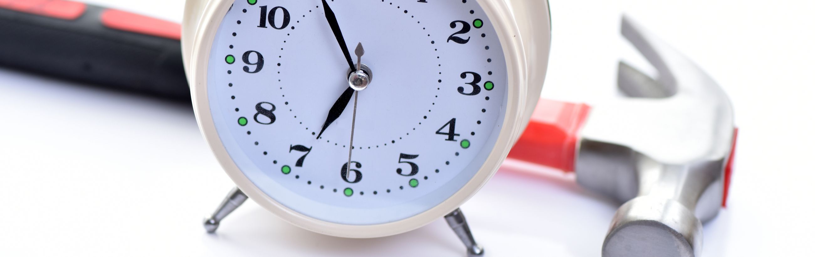 Close-up of a clock face with green dots on some numbers, placed on a white background. Next to the clock, a red and black hammer is partially visible, suggesting tools and time measurement.