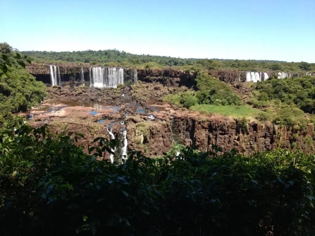 A lush panoramic view of multiple waterfalls cascading over dark basalt cliffs surrounded by vibrant green forests under a clear blue sky.