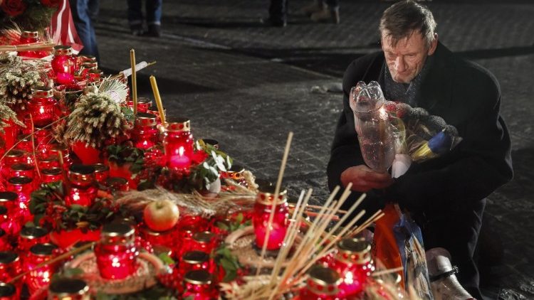 An elderly man touches a makeshift shrine at night, filled with red candle lanterns, incense, and wintry natural offerings like pine cones and sprigs. An evocative scene reflecting traditions that may align with cultural observances under the stars.