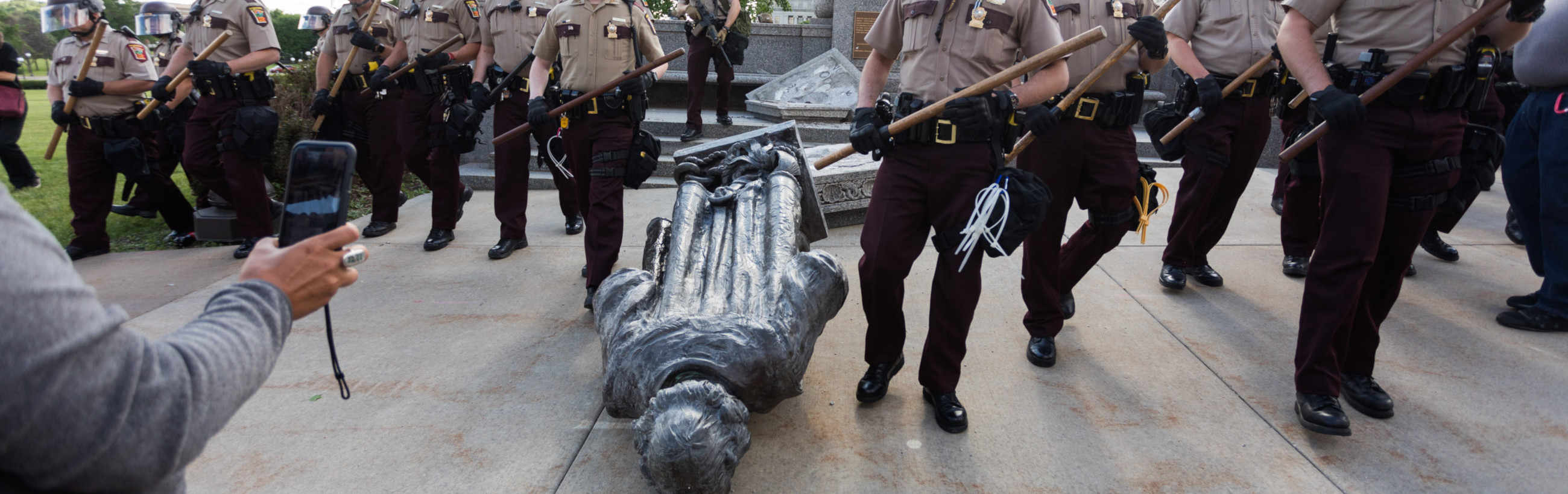 A sculptural representation resembling a figure traditionally associated with the celestial constellation known in biblical terms as Mazzaroth is surrounded by uniformed law enforcement officers carrying batons, with a crowd observing and capturing the event on mobile devices.