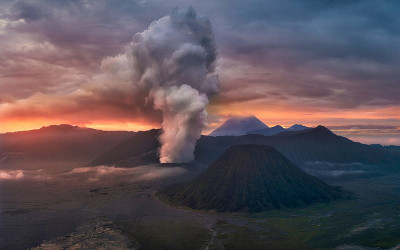 A volcanic eruption spews ash and smoke into the early evening sky, painted with hues of orange and blue. Surrounding the volcano are rugged terrains and distant mountains under a vast sky showing the early signs of the Mazzaroth.