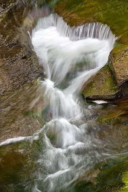 A cascade of water flows smoothly over moss-covered rocks in a forest creek, showcasing the dynamic and natural fluidity of a mountain stream.