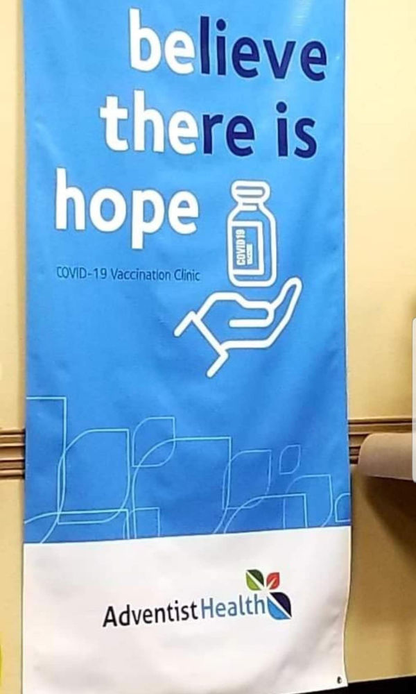 A promotional banner in blue featuring the phrase "believe there is hope" in white letters at the top. Below this, an iconographic representation shows a human hand holding a vaccine vial, labelled “COVID-19 Vaccination Clinic.” The bottom section of the banner carries the logo of Adventist Health.