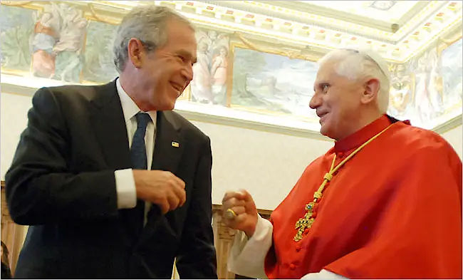 Two elderly men, one in a dark suit and the other in a bright red garment, are sharing a joyful moment with laughter in a lavishly decorated room. Frescoes adorn the walls behind them.