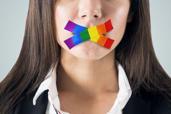 Close-up of a young woman with a piece of multicolored tape over her mouth, symbolizing muted speech. She wears a dark jacket and white shirt, looking directly into the camera.