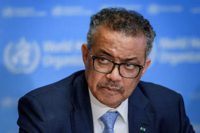 A man with a serious expression, wearing glasses and a dark suit, sits in front of a blue backdrop adorned with a white emblem.