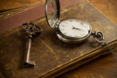 An antique pocket watch with its lid open, resting on an aged book next to an old-fashioned key, all lying on a wooden surface.