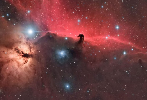 A cosmic vista showcasing a nebula with nebulous red and blue hues surrounding luminous stars. A pillar-like formation resembling a horse's head emerges prominently against a backdrop displaying the vast expanse of the universe.