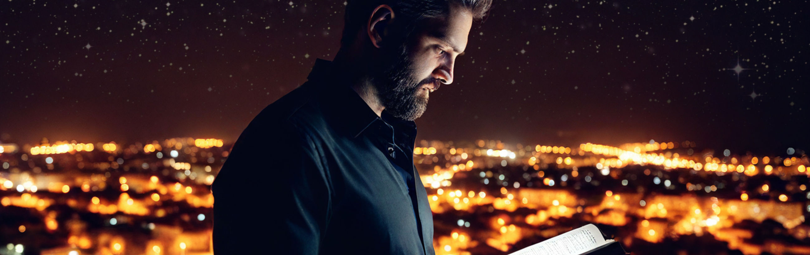 A man with a beard looks at a book, bathed in soft light under a starry sky, with a brightly lit cityscape extending into the distance beneath the celestial display.