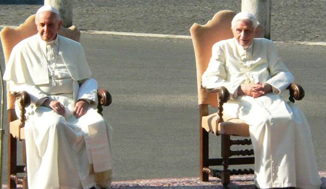 Two elderly men in traditional religious robes sitting on ornate wooden chairs in a sunny outdoor setting.