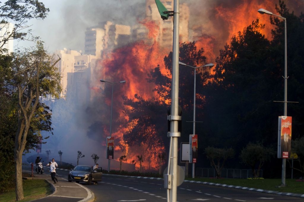 A dramatic urban scene showing a large fire engulfing trees and buildings adjacent to a busy street. Smoke billows as pedestrians and vehicles react near streetlights and signage.